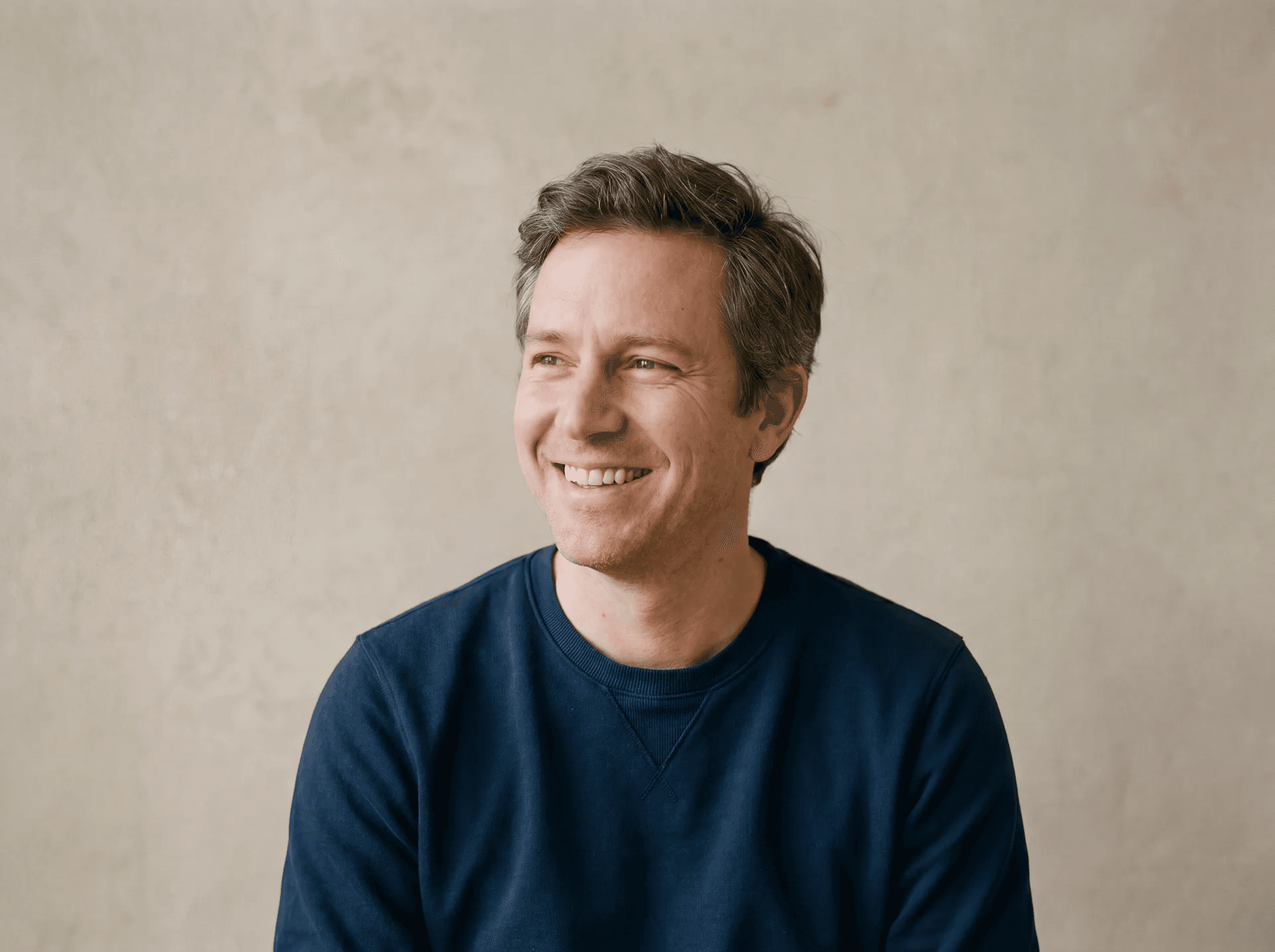 A smiling man in a navy sweater against a warm plaster wall